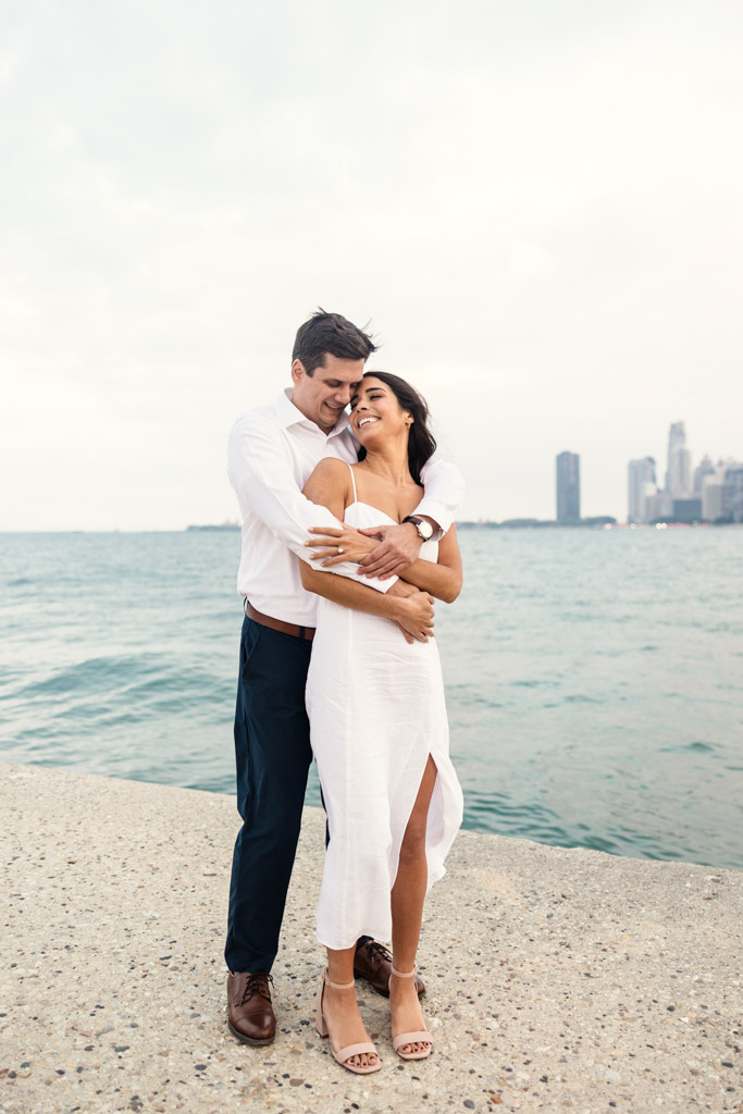 Couple in white clothes smiles and embraces by the water at North Avenue Beach with Chicago skyline in the background during Lincoln Park engagement session