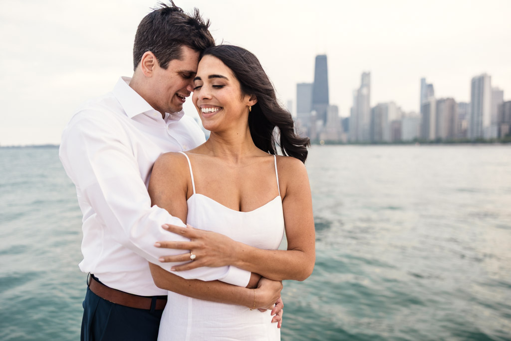 Happy engaged couple holds each other on the shore of Lake Michigan with Chicago skyline in the background