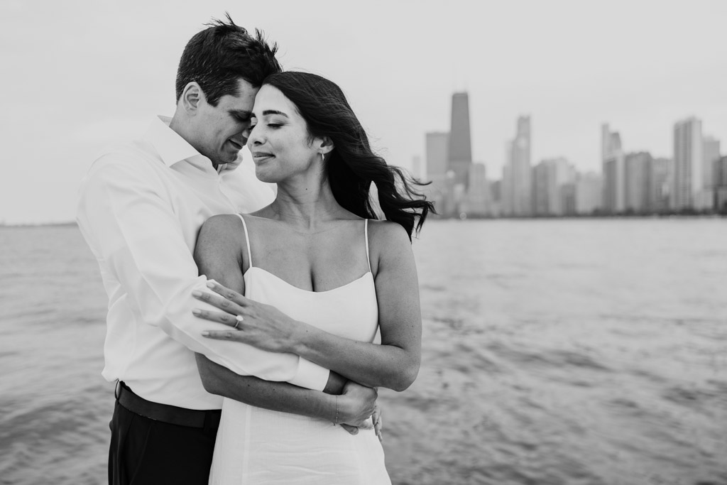 Black adn white photo of engaged couple holding each other near the water in Lincoln Park with Chicago skyline in the background