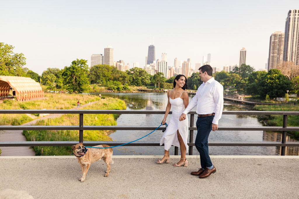 Engaged couple walks their dog on a bridge in Lincoln Park with Chicago skyline in the background