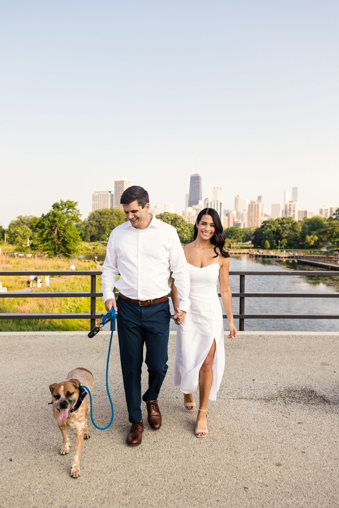 Engaged couple walks their dog on a bridge in Lincoln Park, with downtown Chicago visible in the background