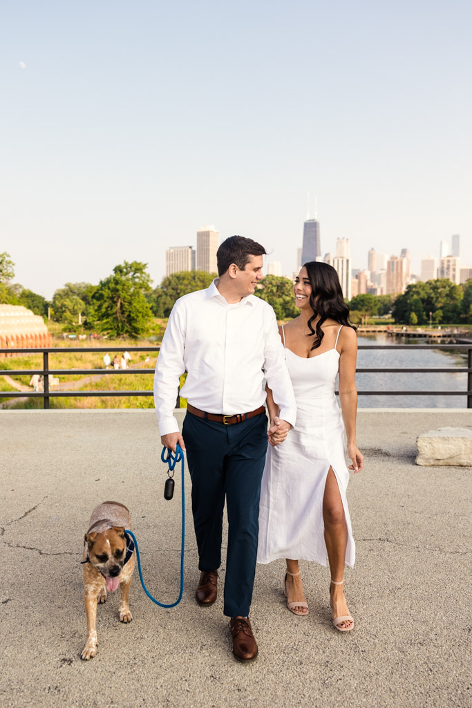 Happy couple walks their dog on a leash on a bridge with Chicago skyline in the background during Lincoln Park engagement session