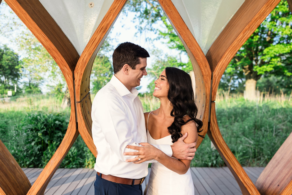 A couple smiles and embraces under the Honeycomb in Lincoln Park during their spring engagement session with greenery in the background.