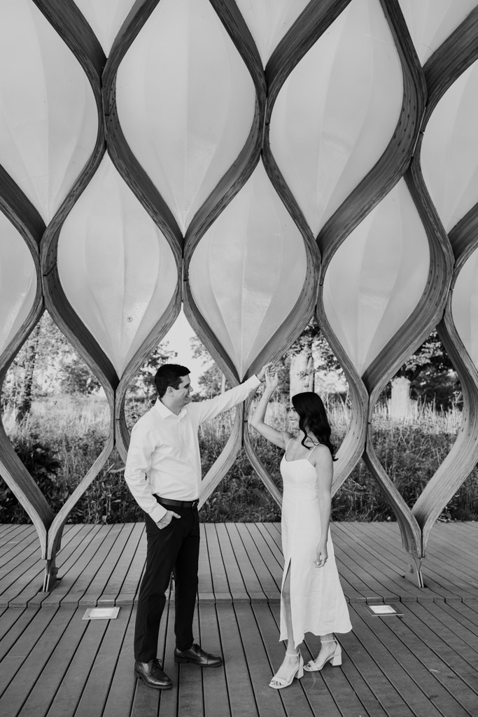 Black and white photo of man twirling his partner as they stand under the Honeycomb in Lincoln Park, holding hands and smiling at each other
