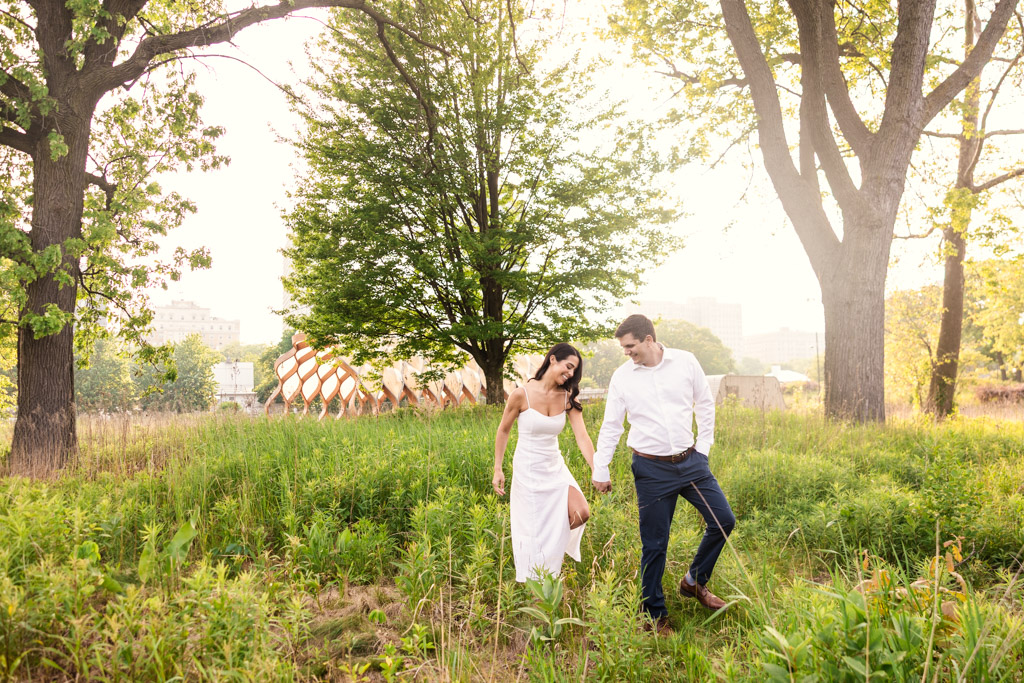 Engaged couple in white clothing walks hand-in-hand through a grassy field in Lincoln Park, with trees and the Honeycomb behind them