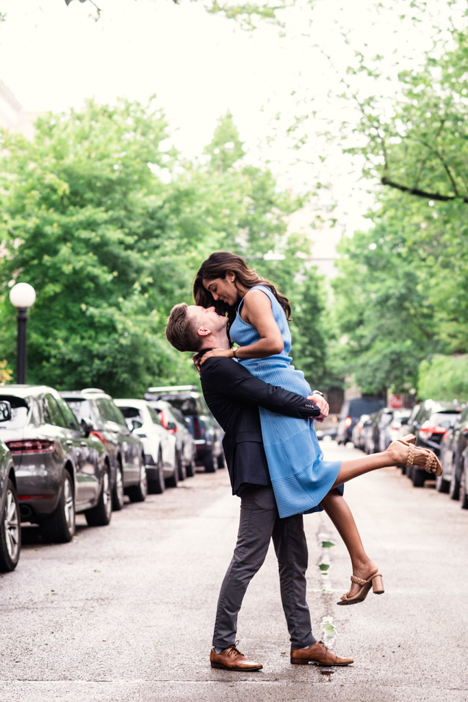 A man lifts a woman in a blue dress on a tree-lined city street during their Chicago summer engagement session