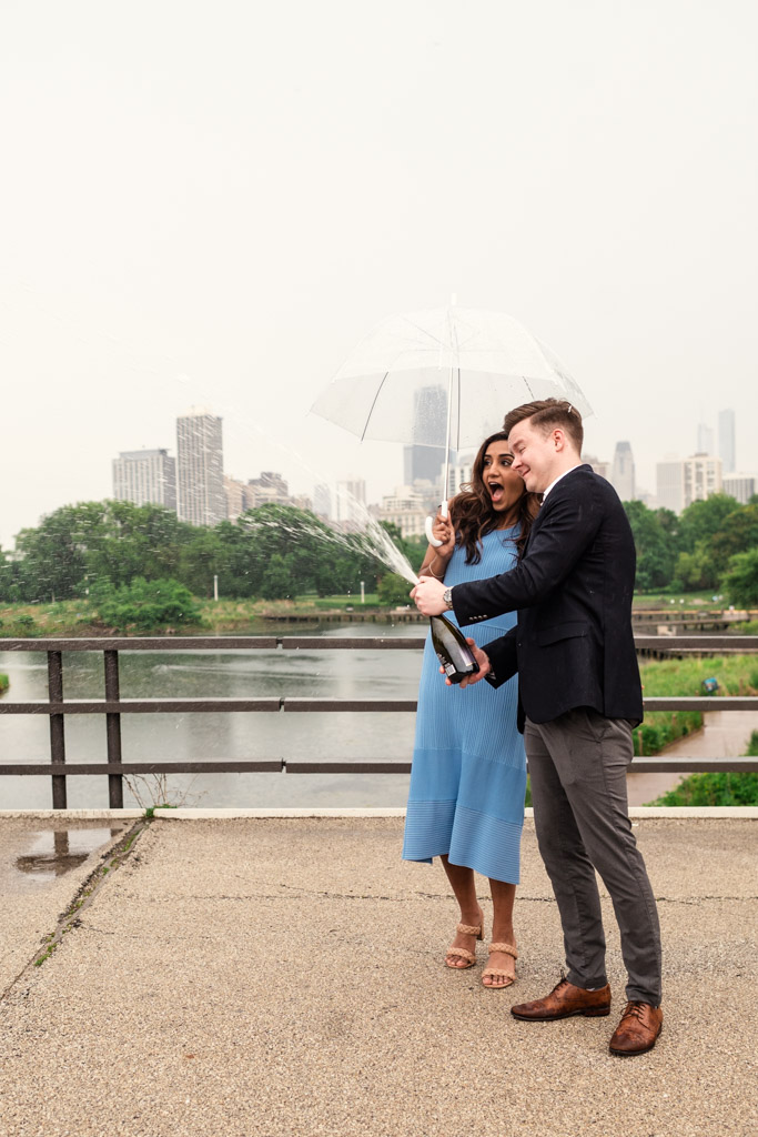 A couple pops a champagne bottle under an umbrella, laughing near a river with city buildings in the background, savoring the unpredictable Chicago weather.