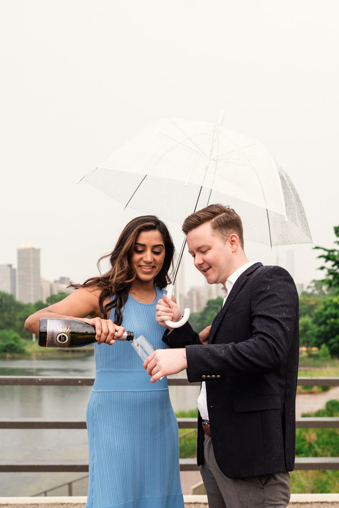 A woman and man share an umbrella and pour champagne in Lincoln Park on a rainy day during their Chicago summer engagement session