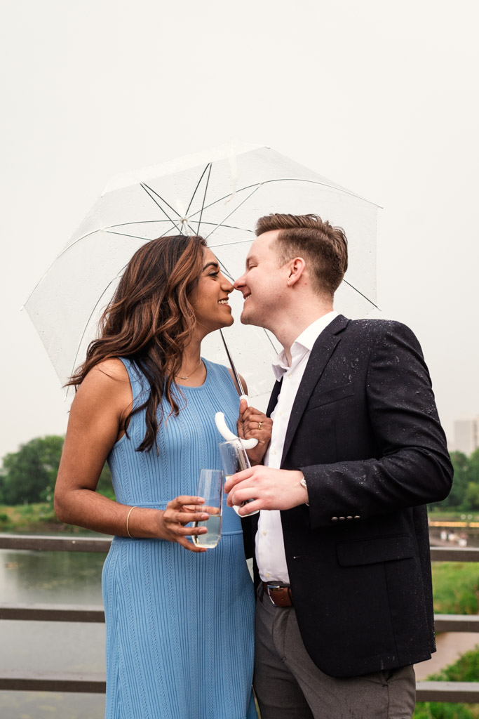 A couple smiles and leans in to kiss under a clear umbrella, holding drinks near a pond in Lincoln Park during their rainy Chicago engagement session