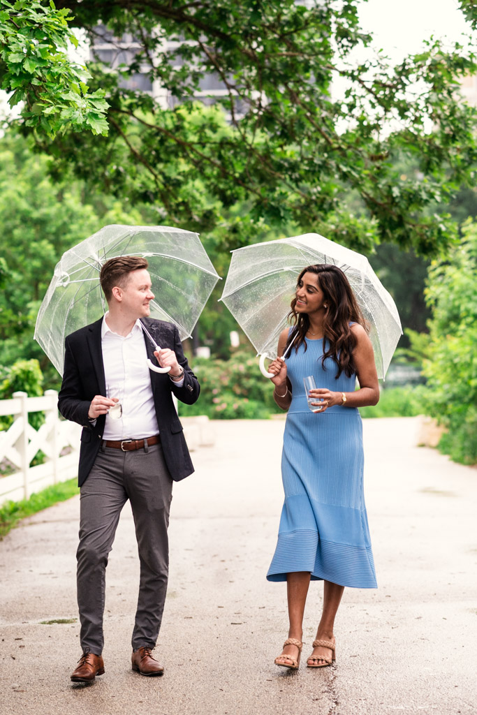 Happy engaged couple walks in Lincoln Park under clear umbrellas, smiling at each other on a tree-lined path