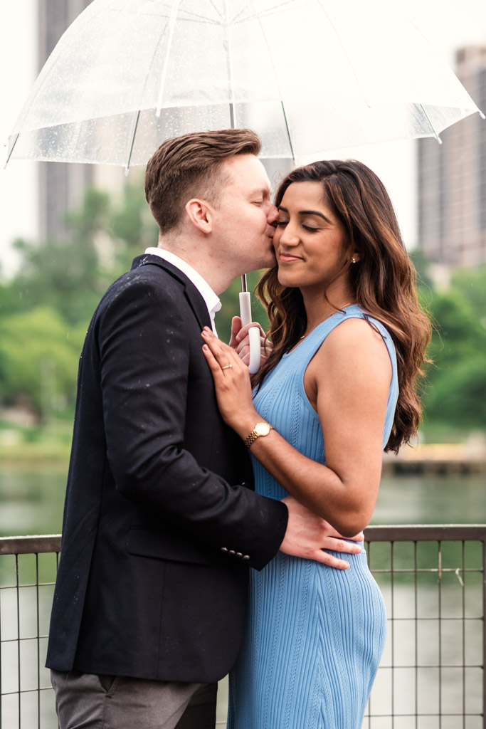 A man kisses his partner on the cheek under a clear umbrella on a Lincoln Park bridge, during their Chicago summer engagement session