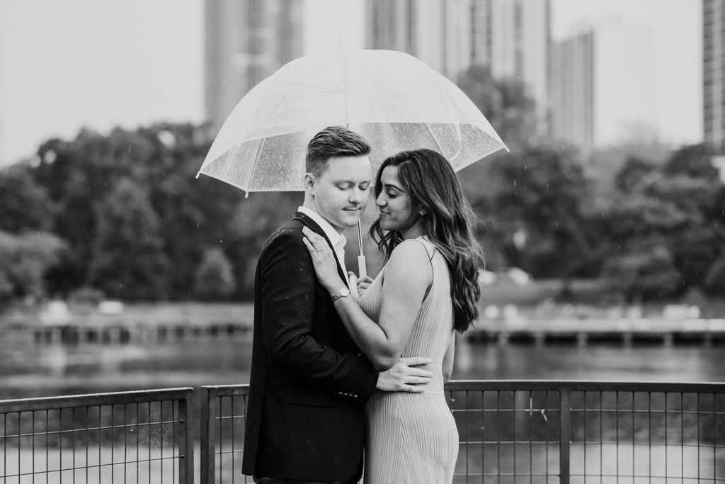 Black and white photo of couple standing under an umbrella on a Lincoln Park bridge in the rain, with city buildings blurred in the background