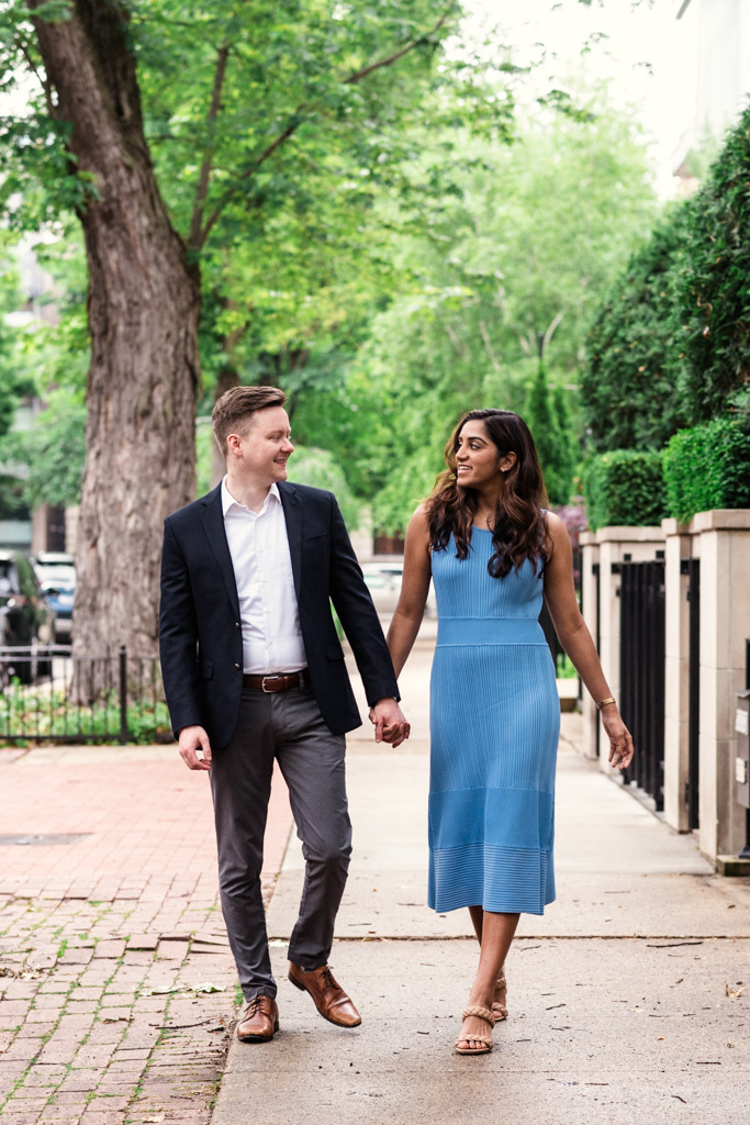 Engaged couple walks hand in hand on a tree-lined sidewalk in Chicago's Lincoln Park neighborhood, smiling at each other