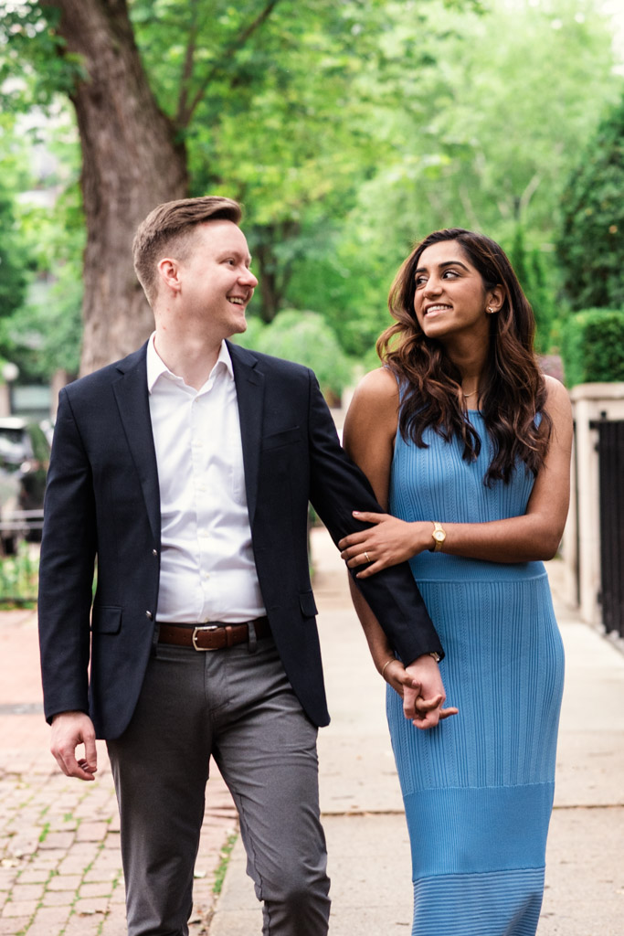 A couple walks outside, smiling and holding hands on a tree-lined sidewalk during their Chicago summer engagement session