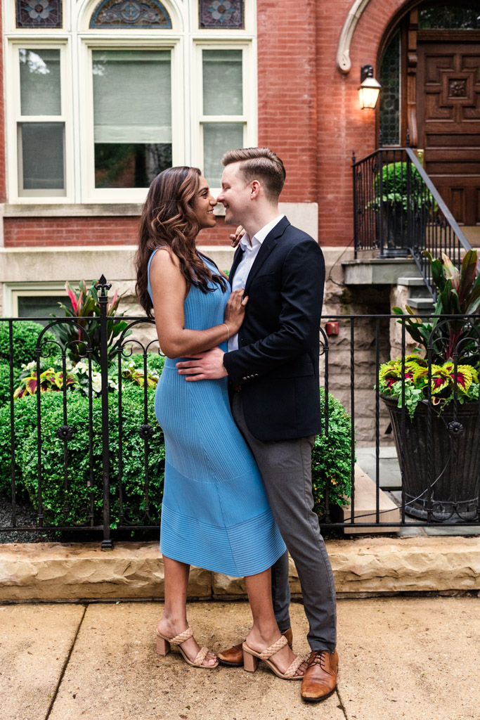 Engaged couple smiles and embraces on a Chicago sidewalk in front of a red brick building and green plants