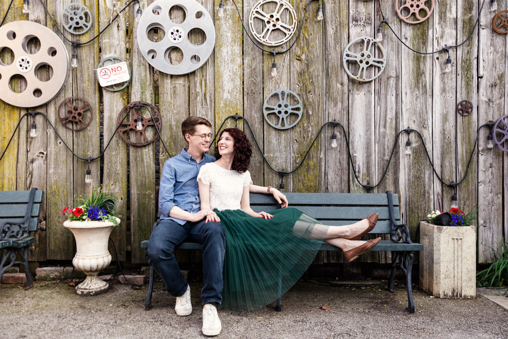 A couple sits smiling on a bench by a wooden fence at the Music Box Theatre Garden, decorated with vintage film reels and potted flowers