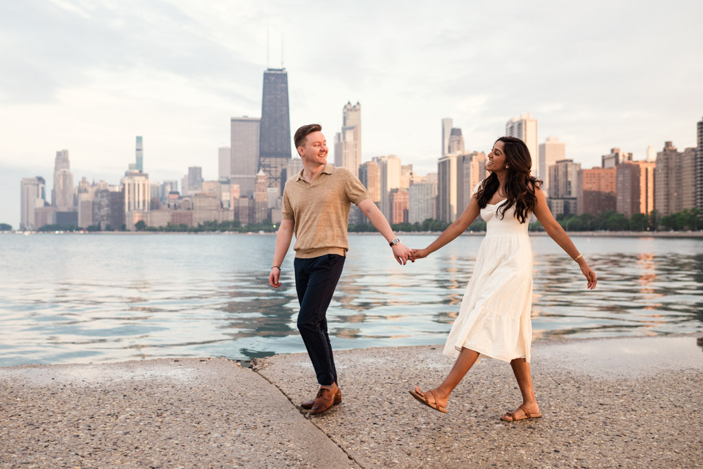 Happy engaged couple holds hands while walking by Lake Michigan at North Avenue Beach with the Chicago city skyline in the background