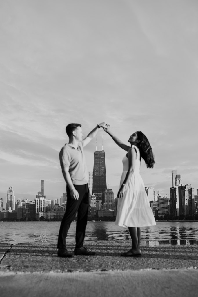Black and white photo of couple dancing by Lake Michigan at North Avenue Beach, with the Chicago skyline in the background during their summer engagement session