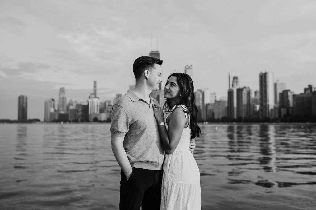 Black and white photo of engaged couple standing by Lake Michigan at North Avenue Beach , smiling at each other, with the city skyline in the background