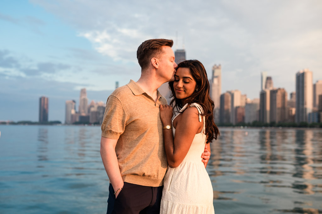 A man kisses a woman’s forehead by the water as the city skyline glows at sunset, capturing a tender moment against the backdrop of beautiful Chicago weather.