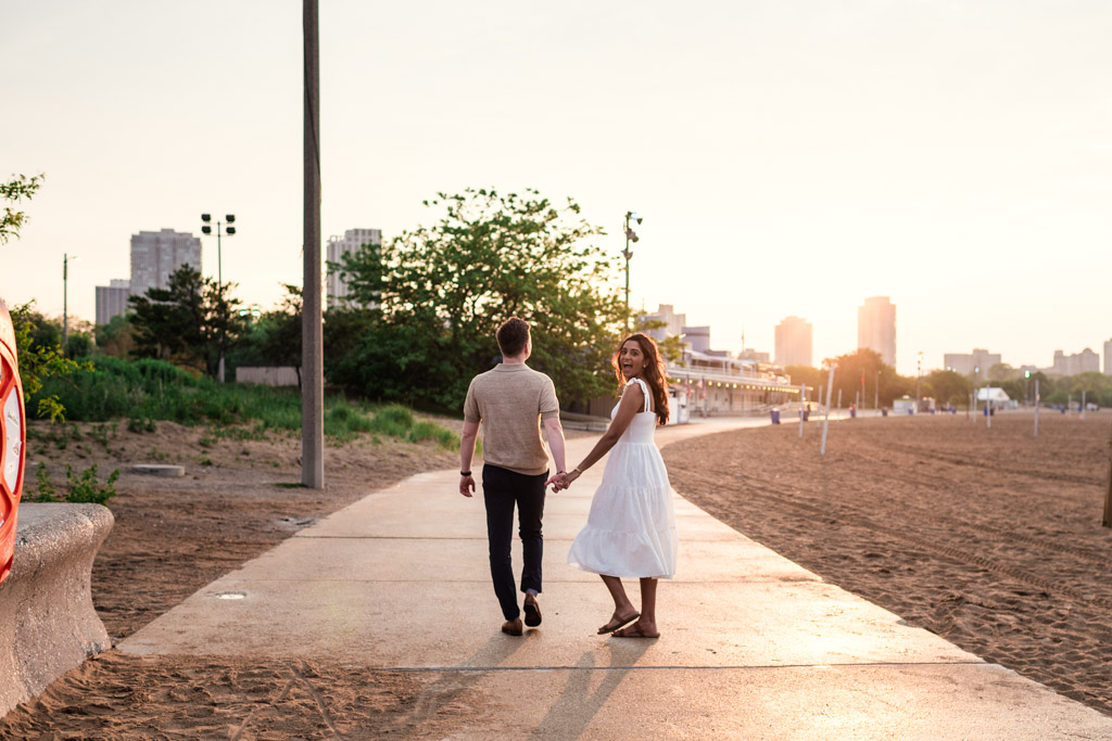A couple holds hands and walks along a North Avenue Beach path at sunset with city buildings in the background