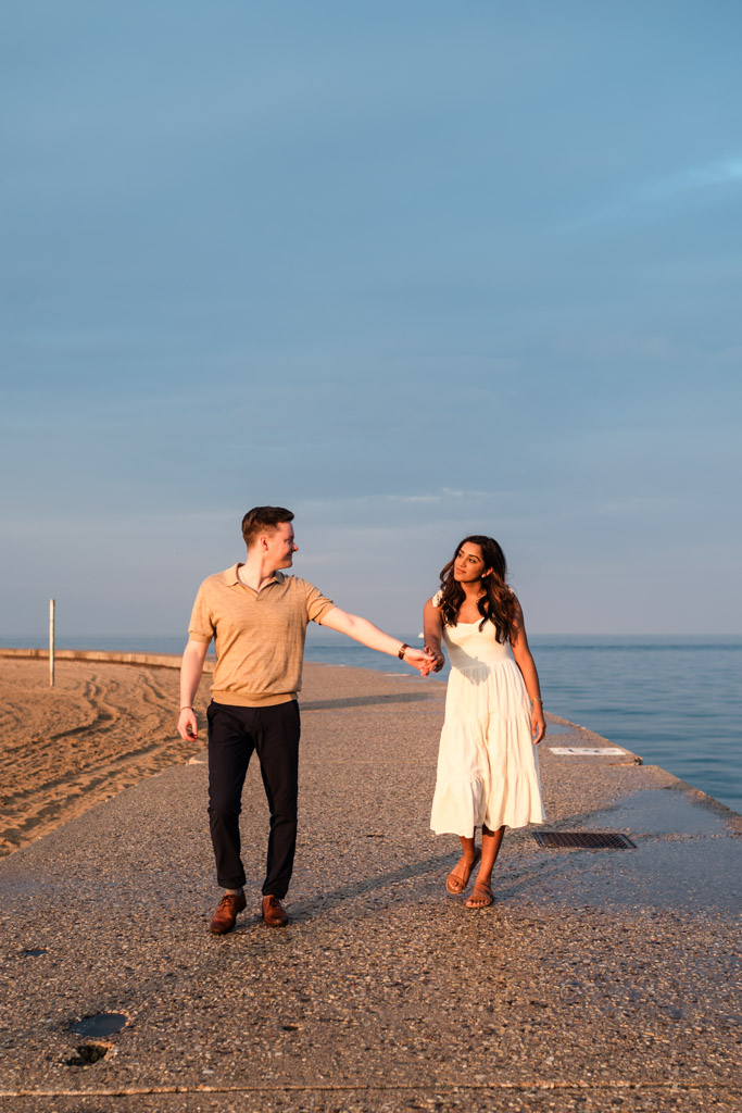 A couple holding hands and walking along a pier at North Avenue Beach at sunset during their Chicago engagement session