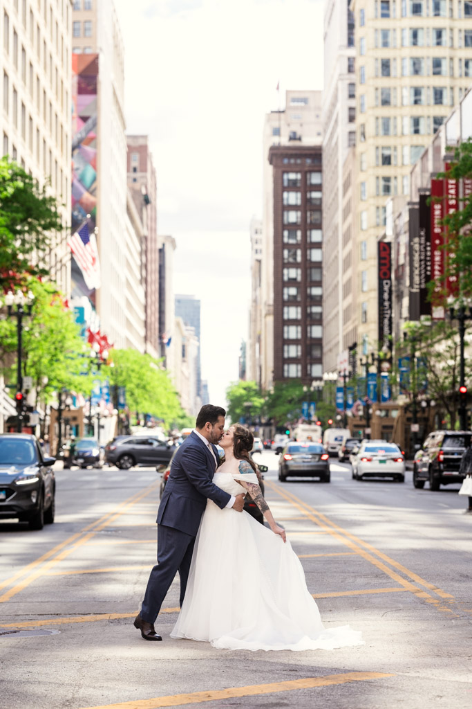 A bride and groom kiss in the middle of a busy city street in downtown Chicago, surrounded by tall buildings and traffic