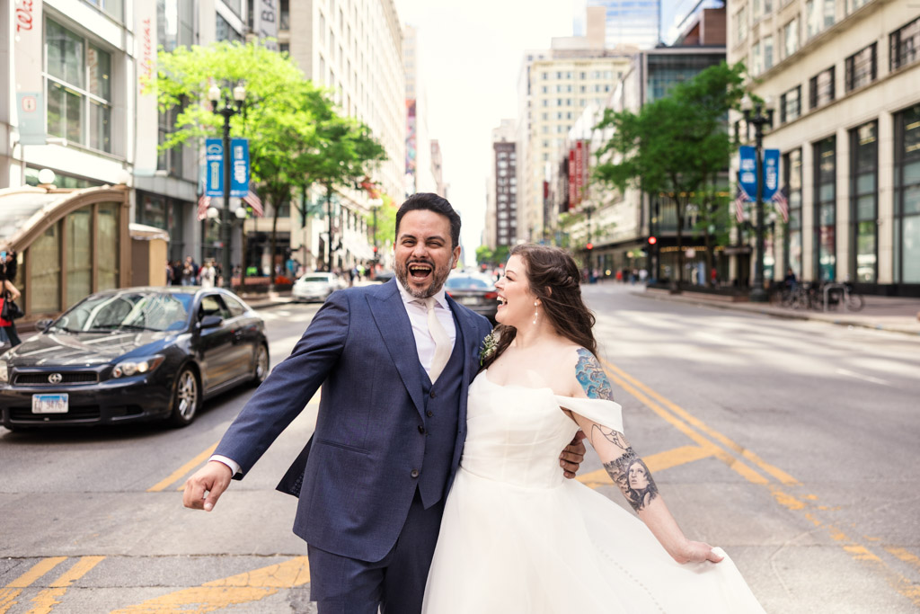 A joyful couple in wedding attire laugh together on a downtown Chicago street, holding hands