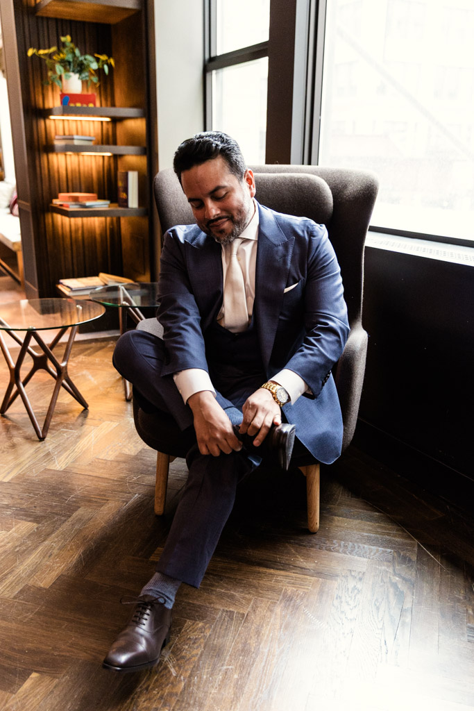 Groom in a blue suit sits in a chair by the window, tying his shoes while getting ready for his wedding at Osteria Via Stato