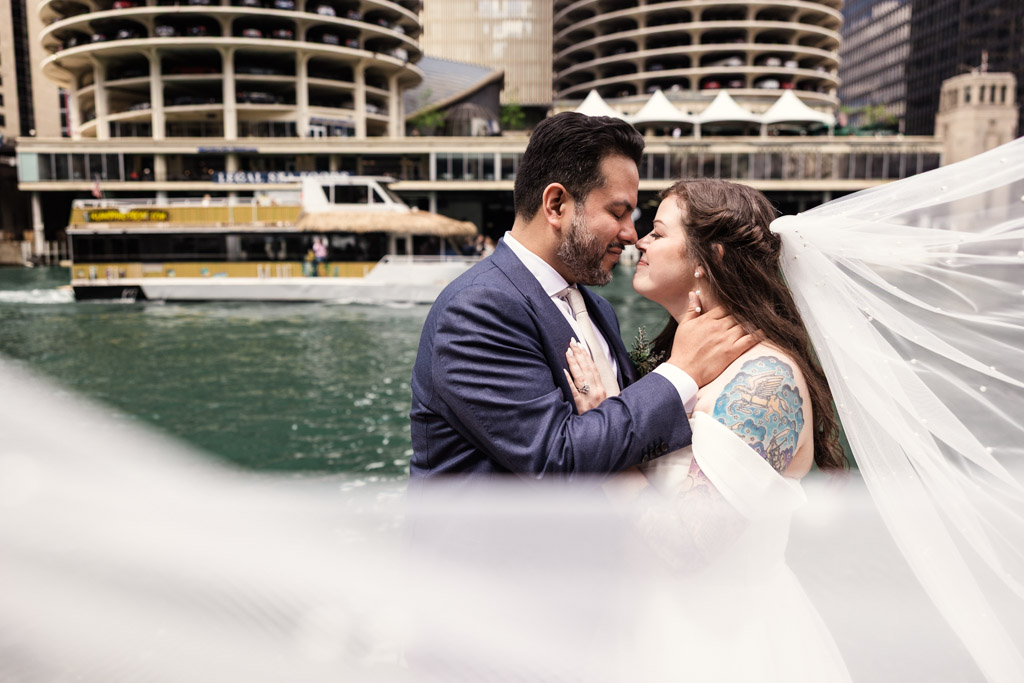 Bride and groom embrace and smile beside the Chicago River, with the bride's veil floating in the wind