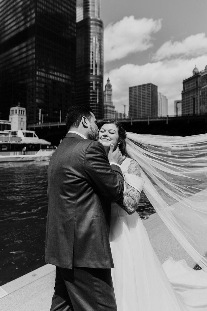 A groom kisses his smiling bride’s face by the Chicago River, city skyscrapers and boats in the background