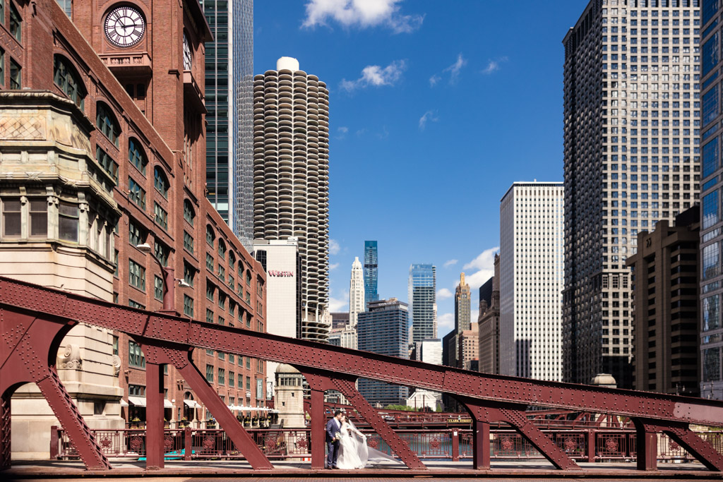 A couple in wedding attire stands on the LaSalle Street Bridge with tall Chicago buildings in the background under a blue sky