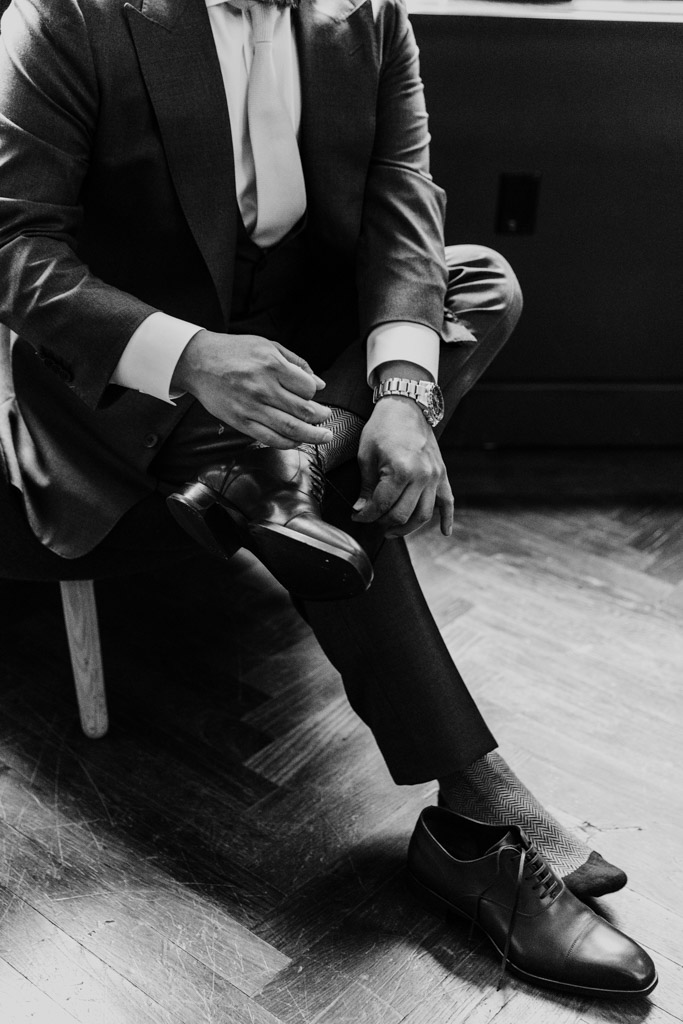 Black and white photo of groom in a suit tying his shoes while getting ready at Virgin Hotels Chicago before his Osteria Via Stato wedding celebration