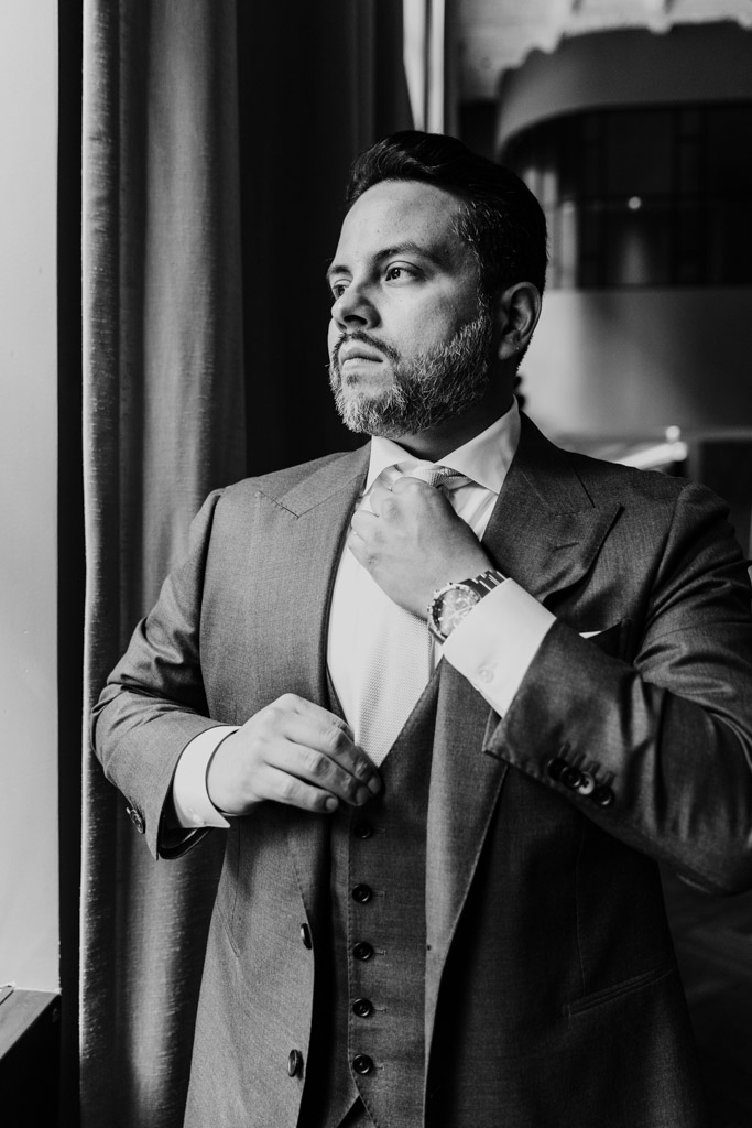 Black and white photo of groom an in a suit adjusting his tie while looking out a window at Virgin Hotels Chicago