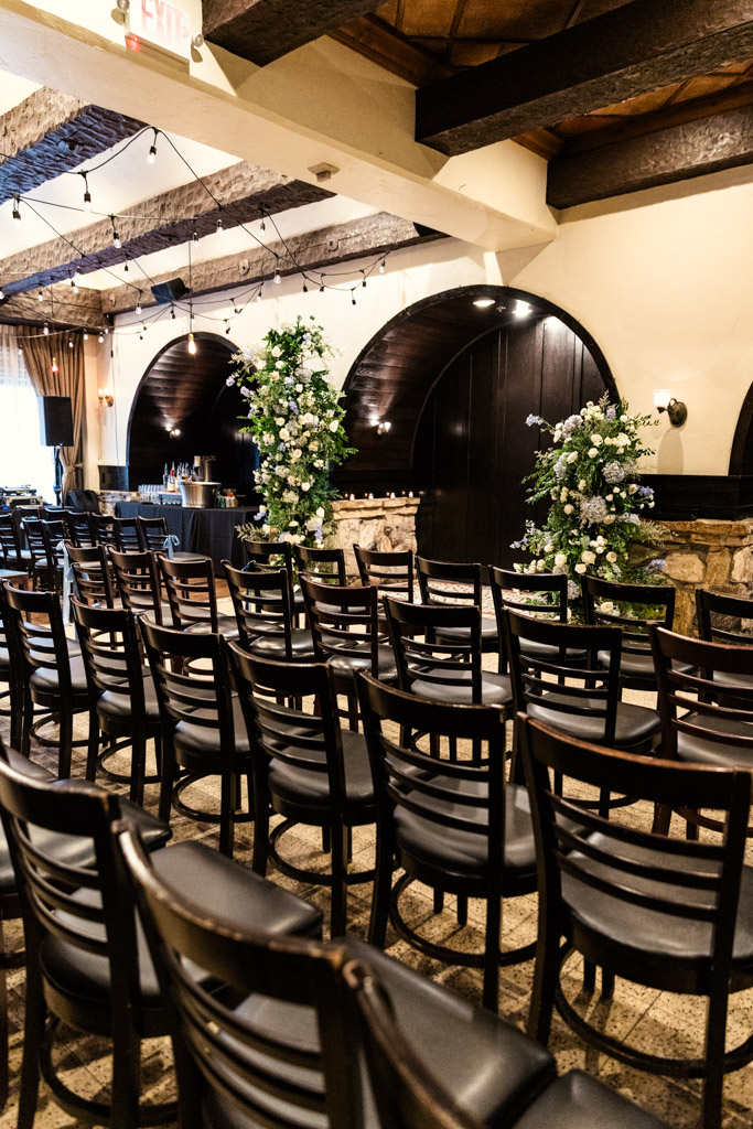 Rows of black chairs face a decorated indoor archway with white flowers and greenery in Osteria Via Stato's wedding ceremony space