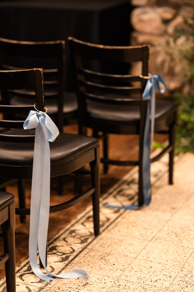 Black chairs lined up with light blue ribbons tied to the backs, indoors on a patterned tile floor at Osteria Via Stato