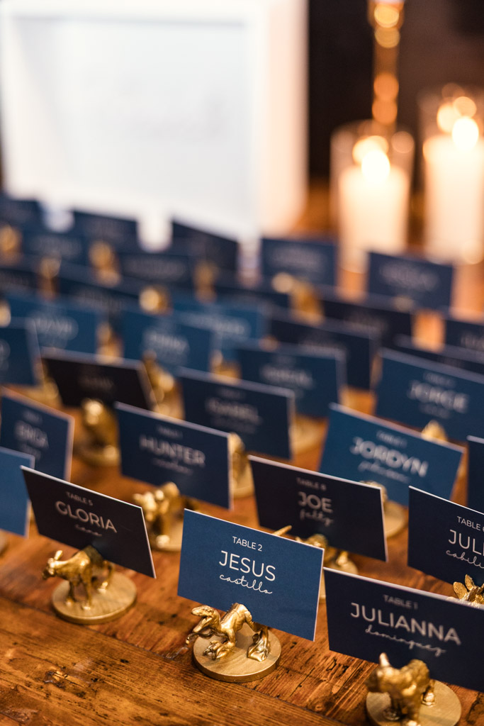 Rows of blue table place cards in gold holders line a wooden table at Osteria Via Stato, with candles softly glowing in the background