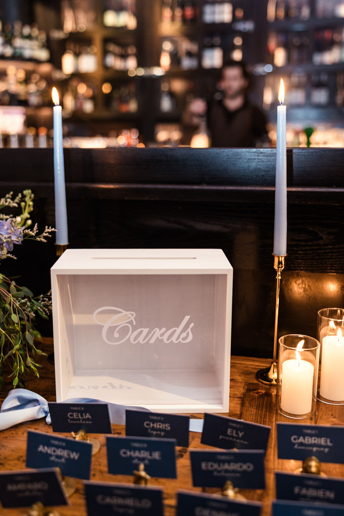 A white card box and lit candles on a wooden table with blue name cards for wedding celebration at Osteria Via Stato