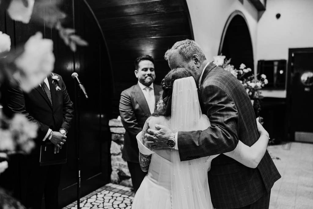A bride in a veil hugs her father during her wedding ceremony at Osteria Via Stato, with the groom smiling nearby