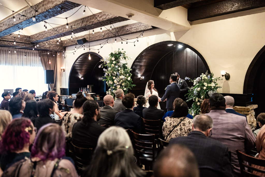 Indoor wedding ceremony at Osteria Via Stato, with guests seated, the couple standing at the the front, and beautiful floral arrangements around them
