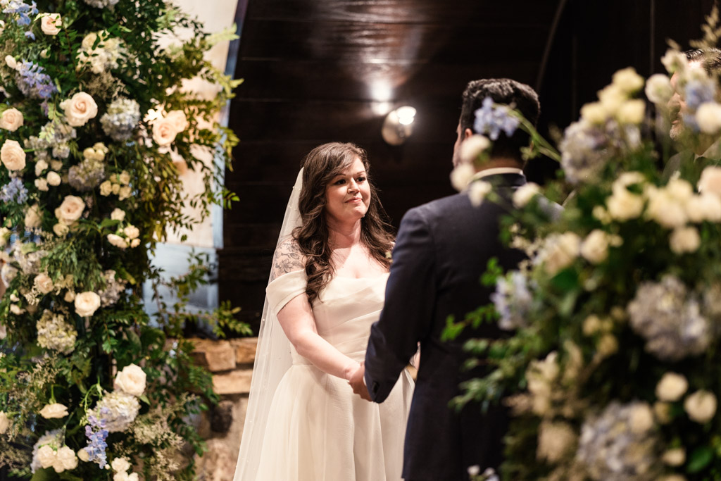 Bride and groom hold hands during their wedding ceremony at Osteria Via Stato, surrounded by large floral arrangements