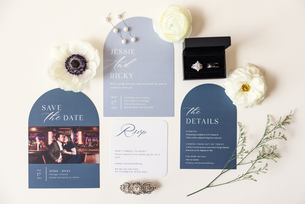 Flat lay of blue and white wedding invitations, rings, flowers, and jewelry on a white background for Osteria Via Stato celebration