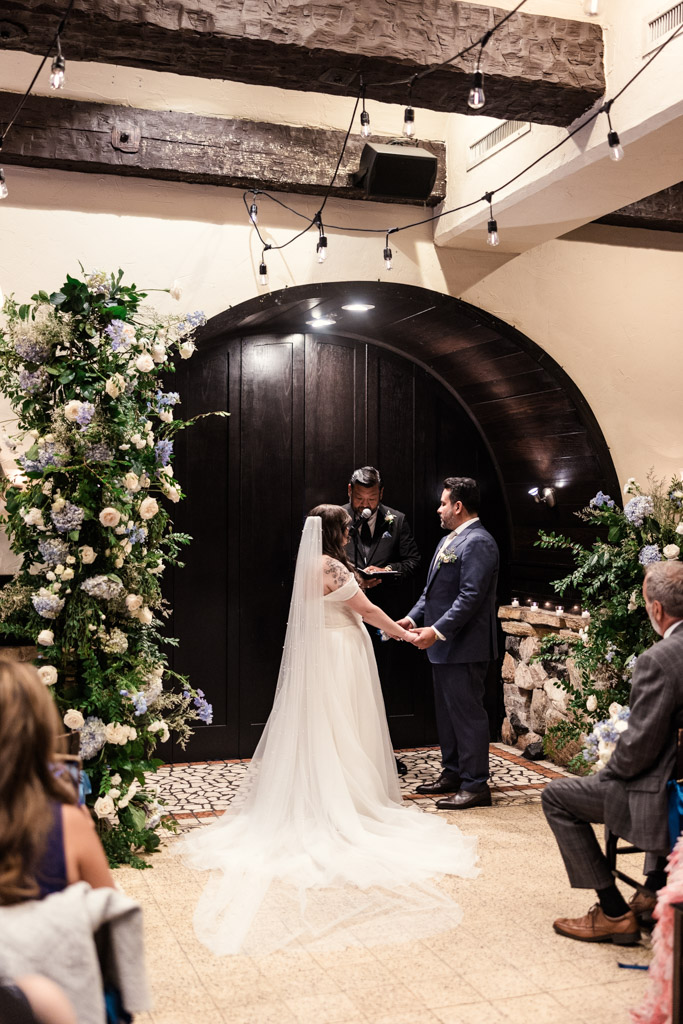 A bride and groom hold hands during their indoor wedding ceremony at Osteria Via Stato, standing before an officiant and elegant floral arrangements.