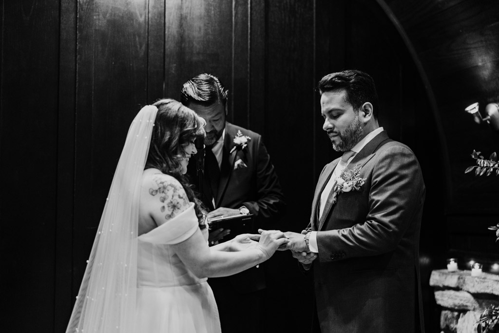 Black and white photo of bride and groom exchanging rings during their wedding ceremony at Osteria Via Stato, with an officiant standing behind them