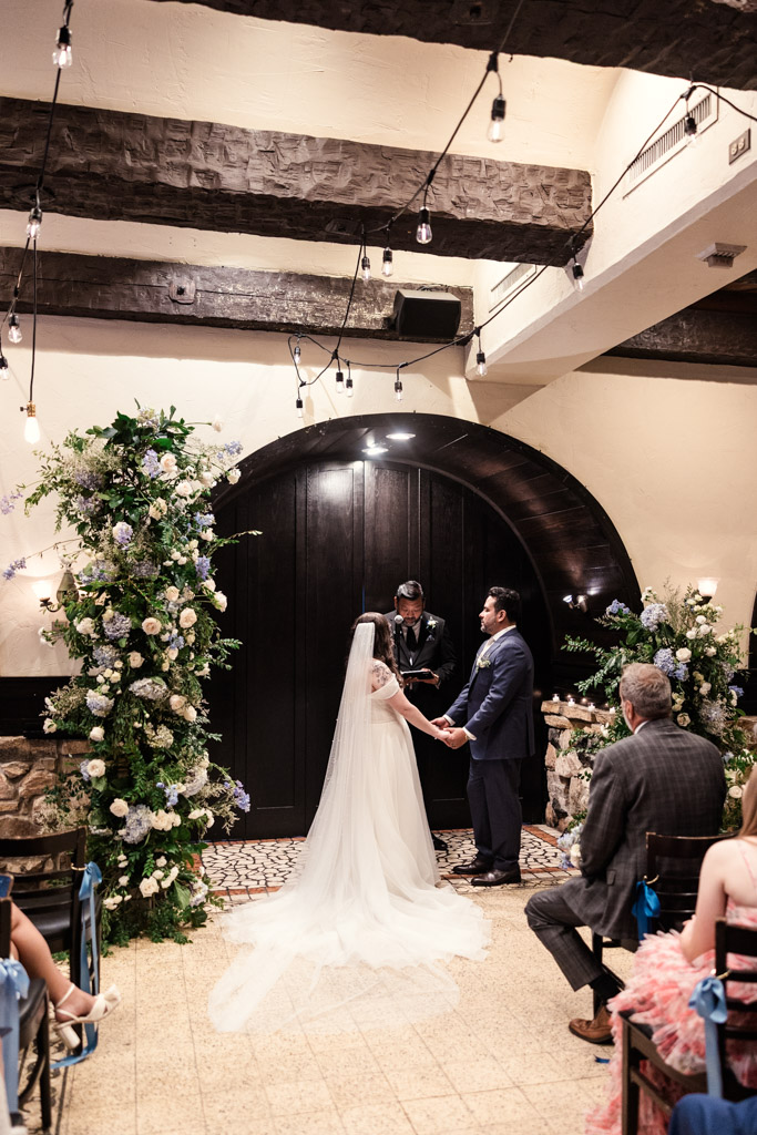 A bride and groom hold hands at the front of their indoor wedding ceremony with floral decorations at Osteria Via Stato