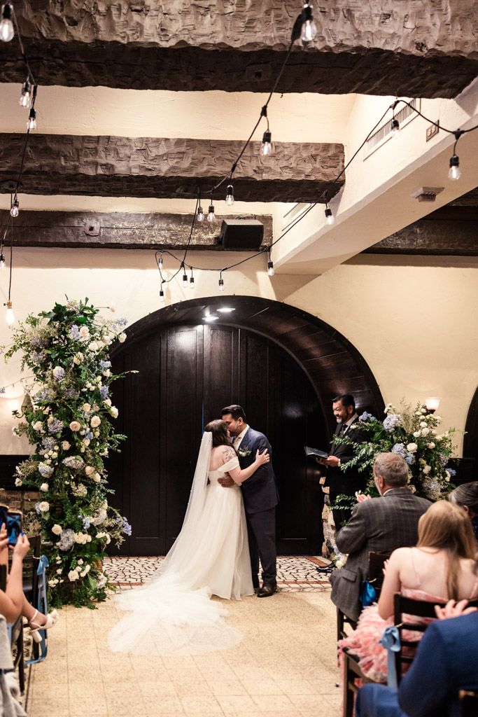 Bride and groom kiss at their Osteria Via Stato indoor wedding ceremony, surrounded by floral arrangements and guests