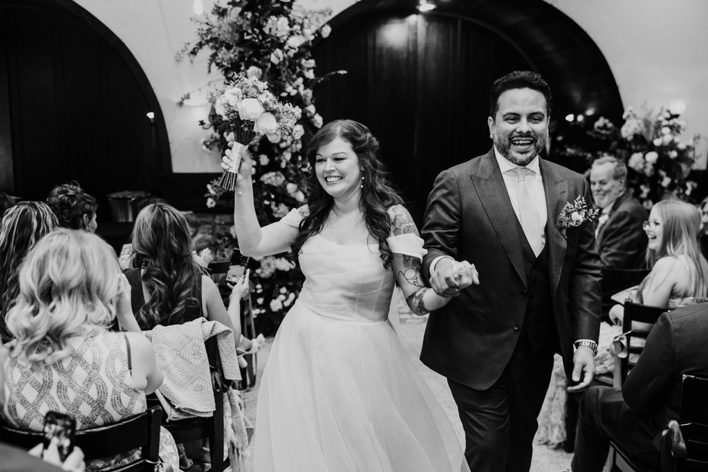 Black and white photo of happy newlyweds walking down the aisle at Osteria Via Stato, holding hands, as guests smile and celebrate