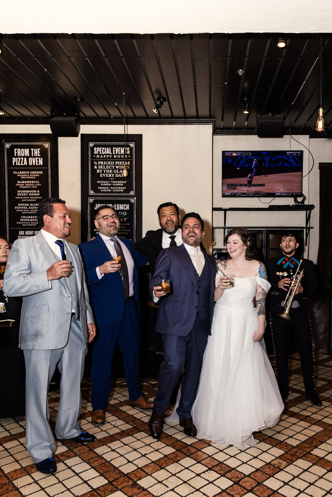 A bride and groom celebrate with drinks at Osteria Via Stato, joined by smiling friends in suits and a mariachi musician