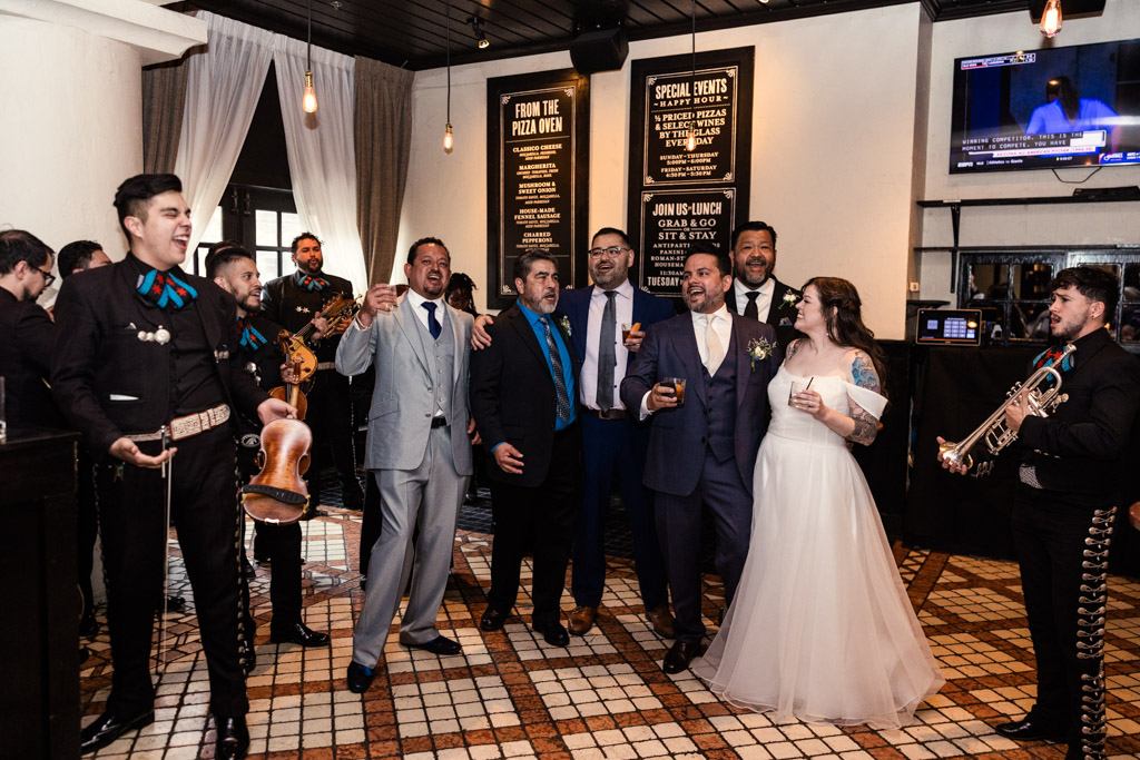 A bride and groom laugh with friends and mariachi musicians during their wedding celebration at Osteria Via Stato