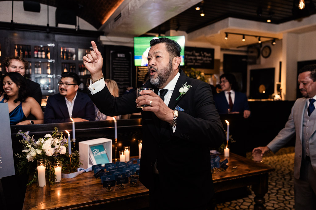Groom's father in a black suit holding a drink and gesturing while singing at a wedding reception at Osteria Via Stato